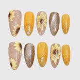 Almond-shaped press-on nails with hand-painted 3D sunflower designs, combining yellow, nude, and brown tones with glitter, gold foil, and transparent details for a warm autumn aesthetic.