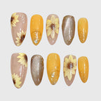 Almond-shaped press-on nails with hand-painted 3D sunflower designs, combining yellow, nude, and brown tones with glitter, gold foil, and transparent details for a warm autumn aesthetic.