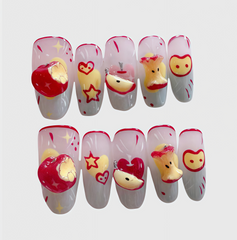 Oval-shaped press-on nails with red apple and fruit core 3D designs, featuring pink and gray gradient background, hearts, stars, and playful cartoon details.